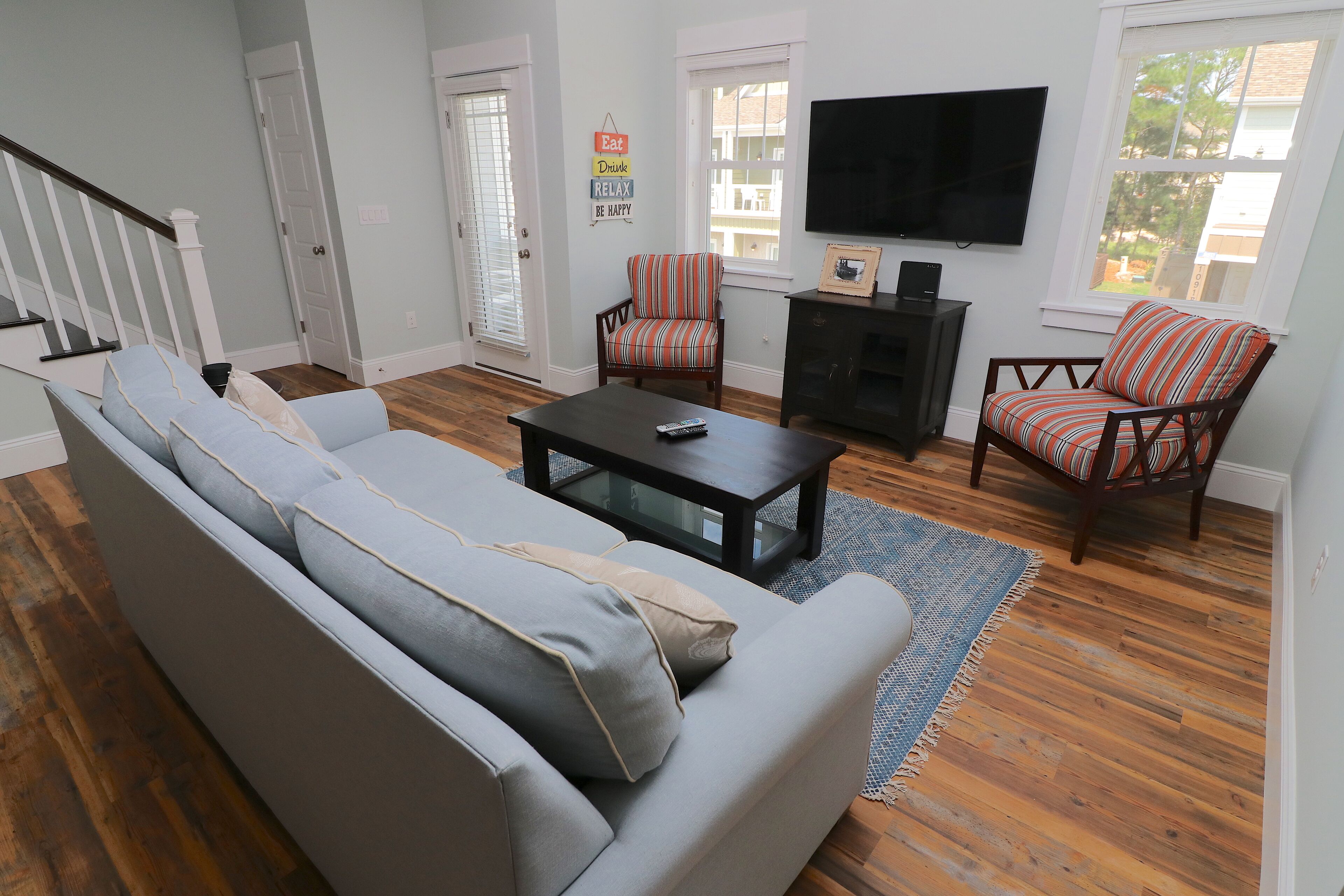 Uma sala de estar com um sofá cinzento, duas poltronas listradas, uma mesa de centro e uma televisão de tela plana fixada na parede.