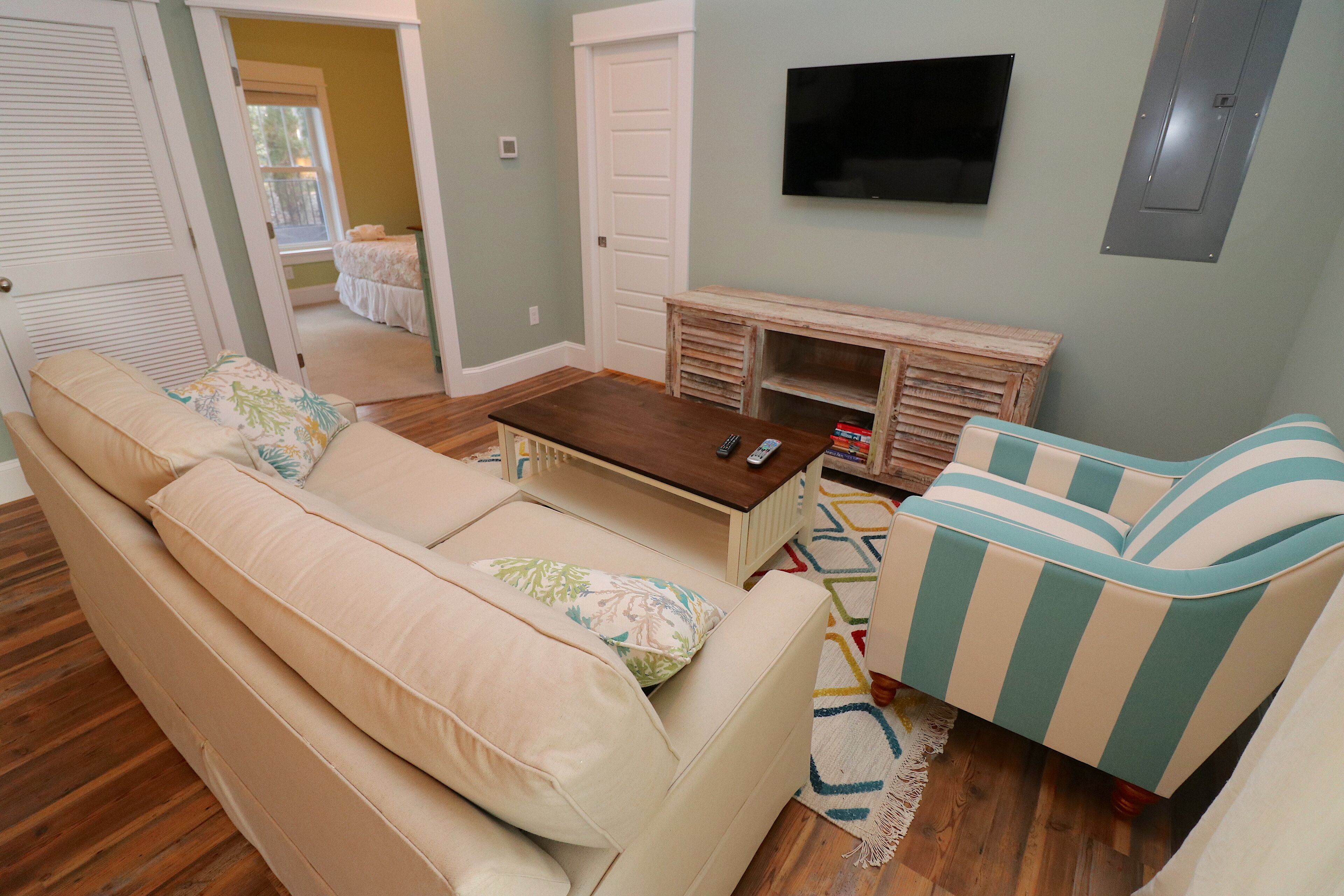Een woonkamer met een beige bank, een gestreepte fauteuil, een houten salontafel en een aan de muur bevestigde televisie.