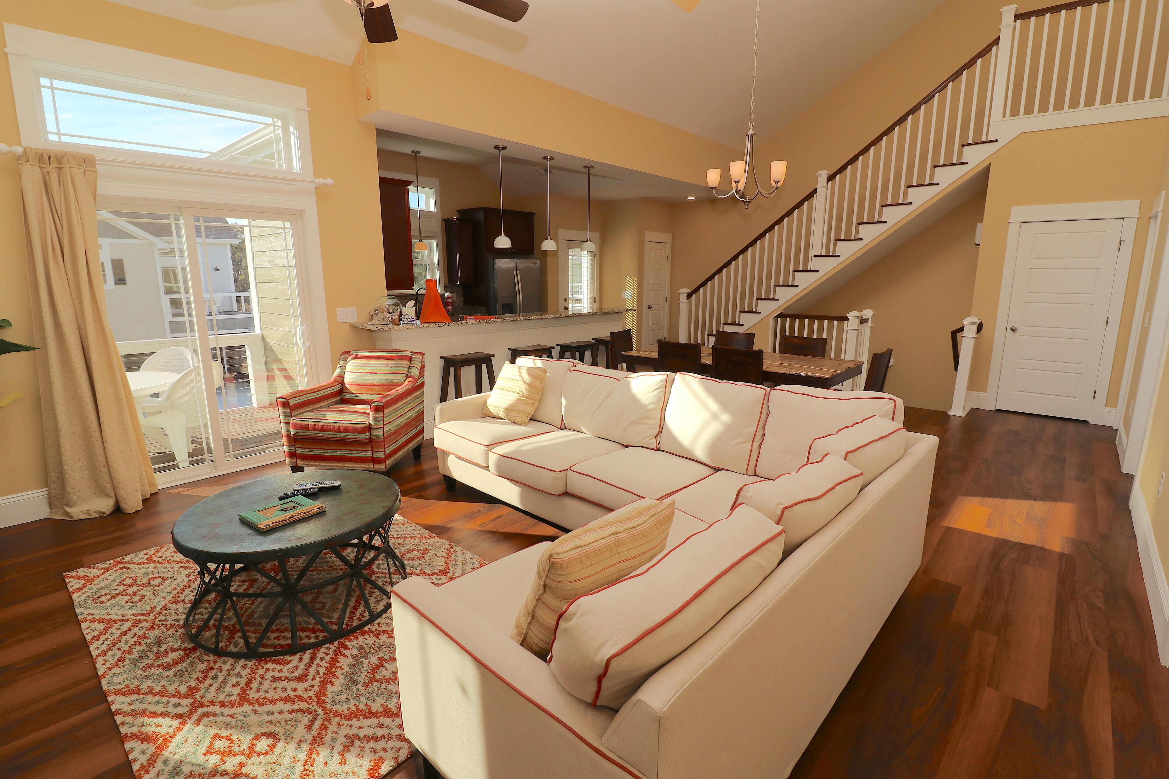 A spacious living room with a large beige sectional sofa, a coffee table, and a dining area with chairs. A staircase leads to an upper level.