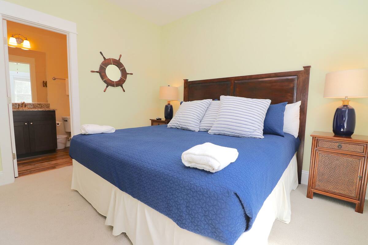 Un lit bien fait, avec une couvre-lit bleue et des rideaux blancs, une tête de lit en bois, deux lampes de chevet et une décoration murale représentant un gouvernail de navire.