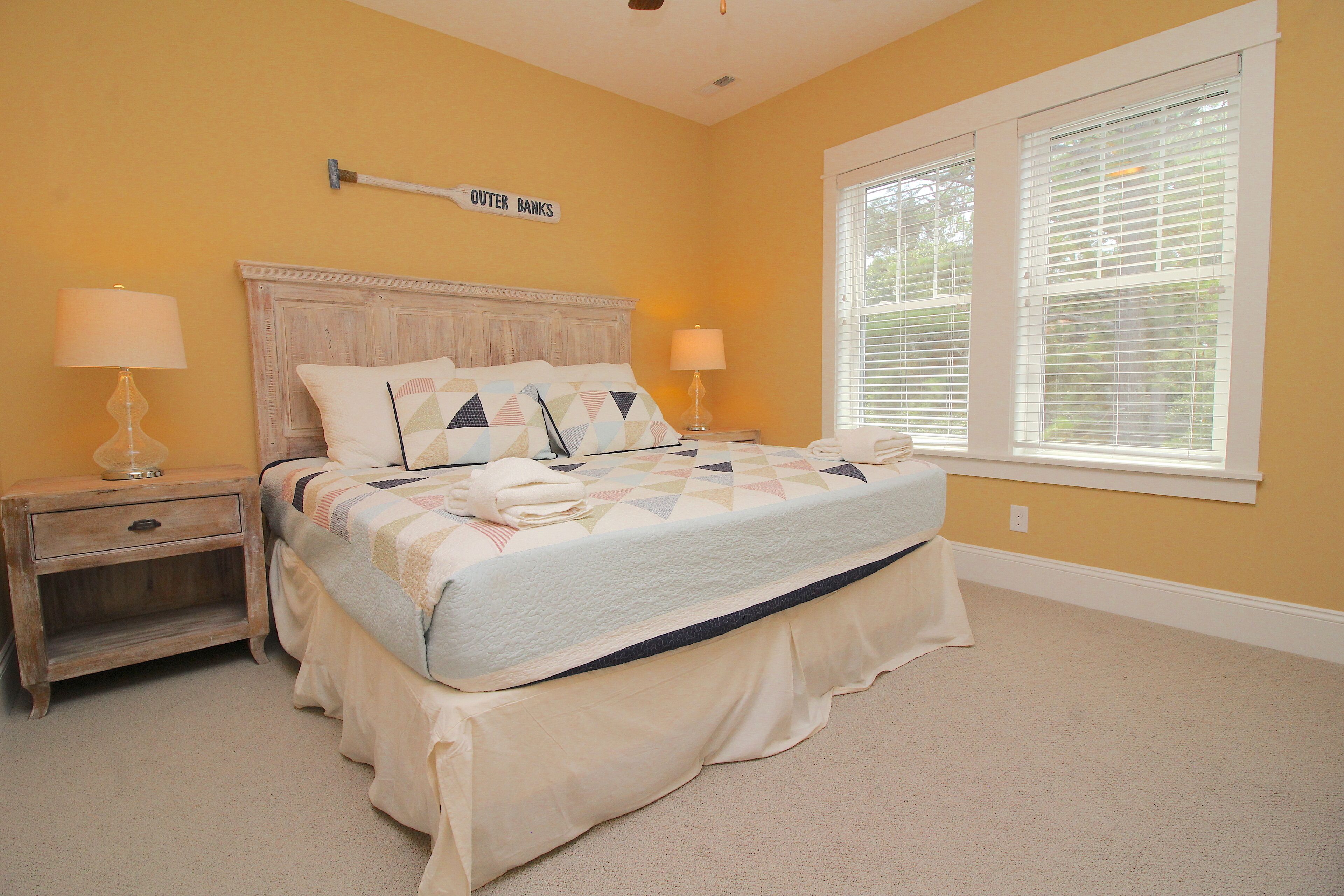 A bedroom with a bed, two nightstands, a wall sign, and a window with blinds.