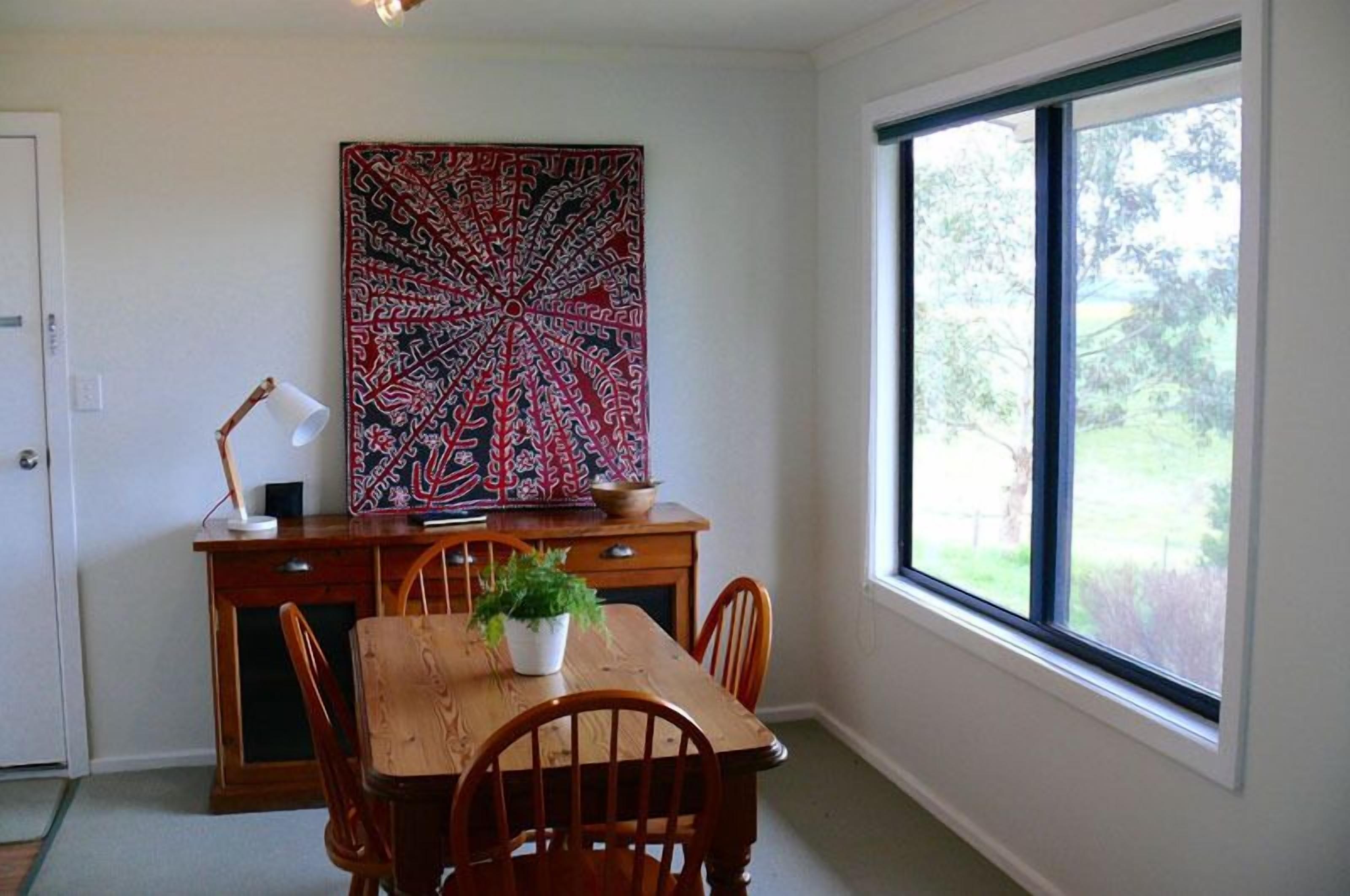 A dining area with a wooden table and chairs, a potted plant, a large abstract painting on the wall, and a window with a view of greenery.