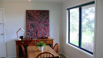 A dining area with a wooden table and chairs, a potted plant, a large abstract painting on the wall, and a window with a view of greenery.