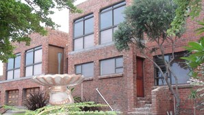A brick building with multiple windows and a small garden area with a decorative stone structure. - Bluewater Beach Accommodation (Gqeberha (Port Elizabeth))