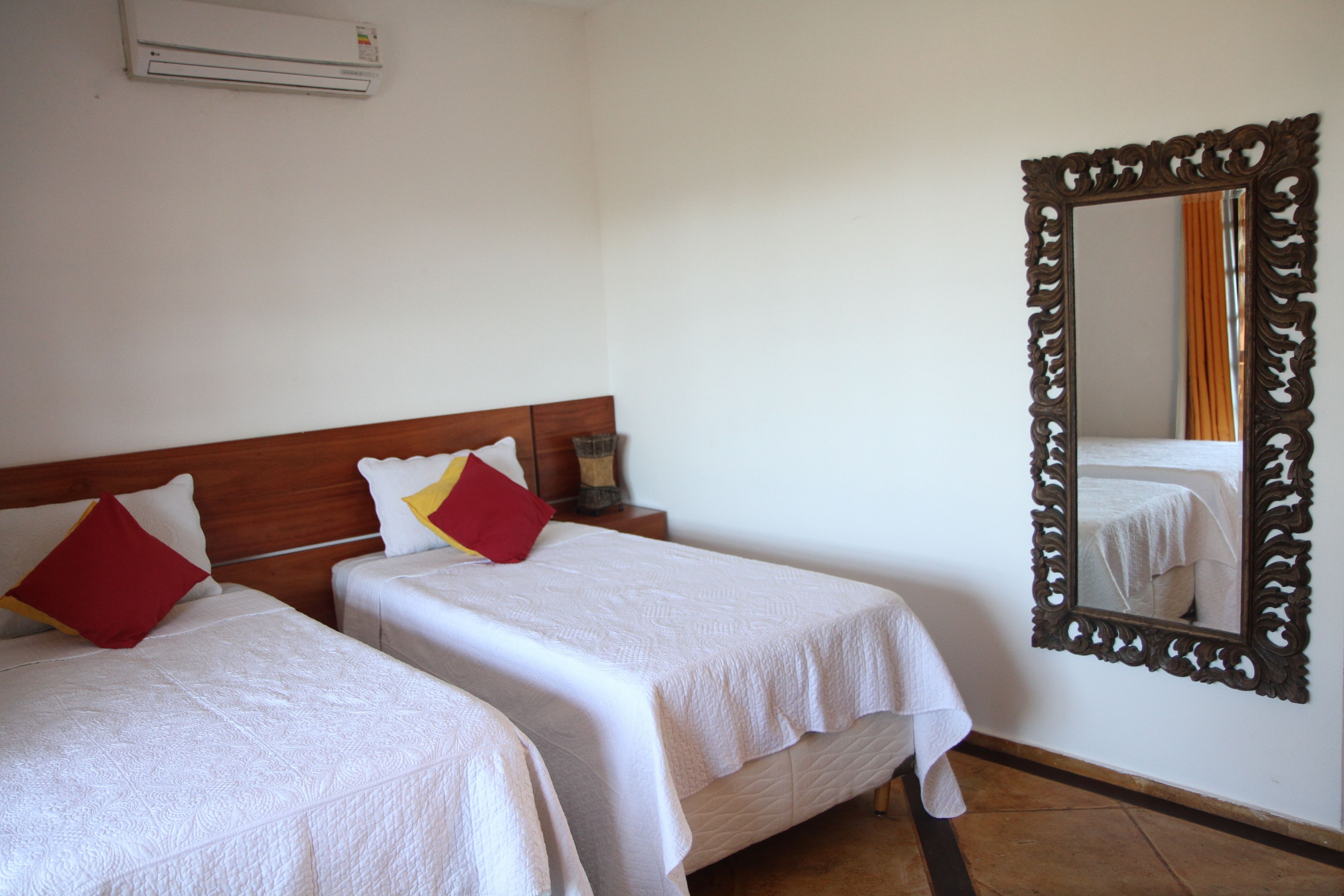 Two single beds with white bedding and red pillows, a wooden headboard, a large ornate mirror, and an air conditioning unit on the wall.