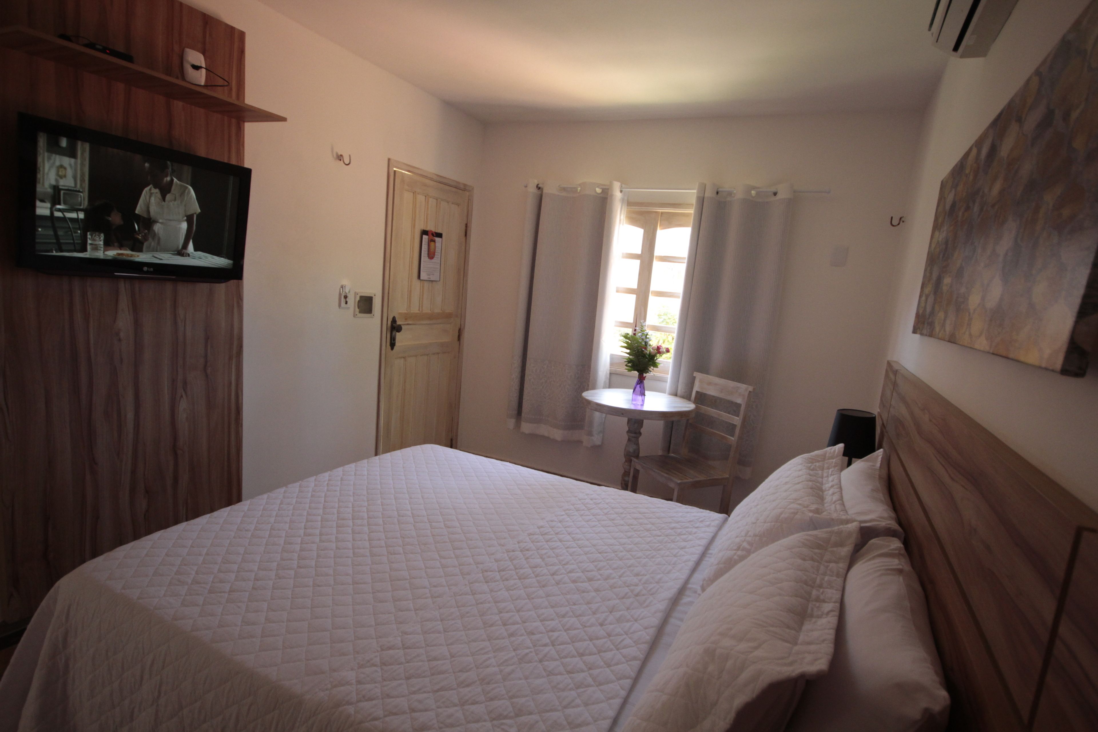 A bedroom with a bed, a TV mounted on a wooden panel, a small table with a vase, and a window with curtains.