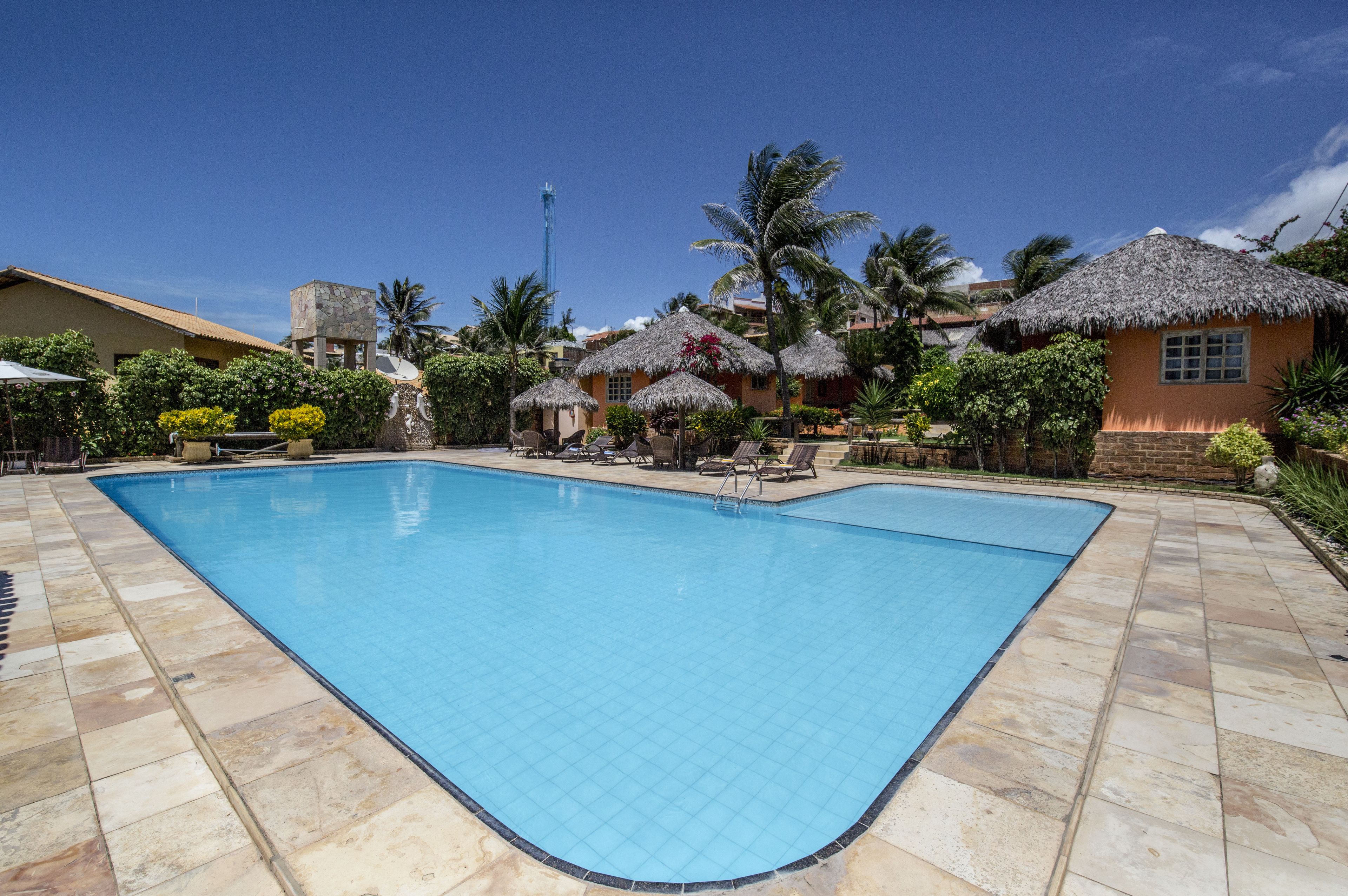 A swimming pool with tiled edges, surrounded by palm trees and thatched-roof huts.