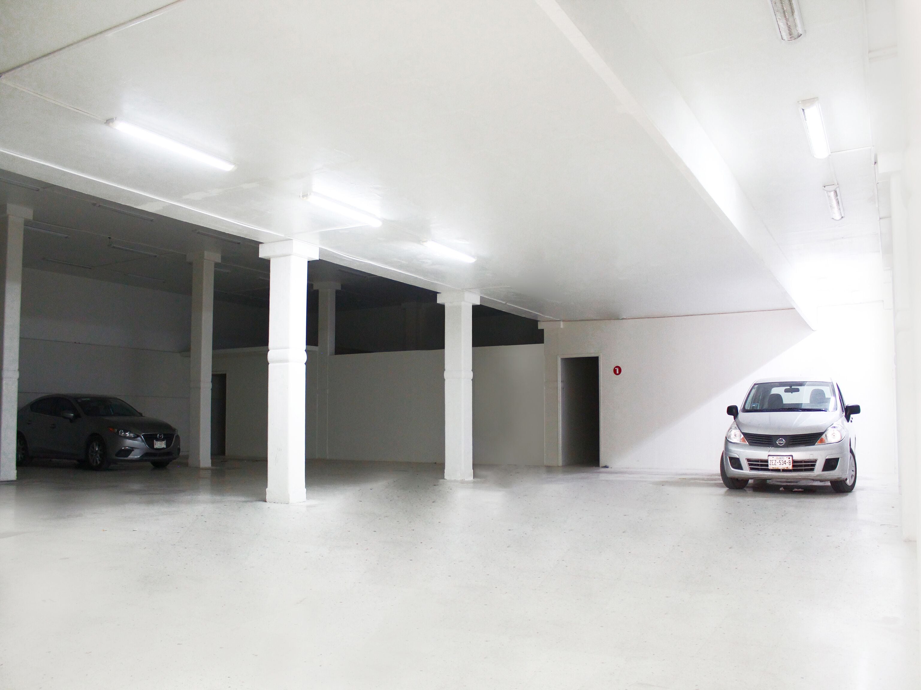 A parking garage with white walls and columns, two cars parked, and a door marked with the number 1.
