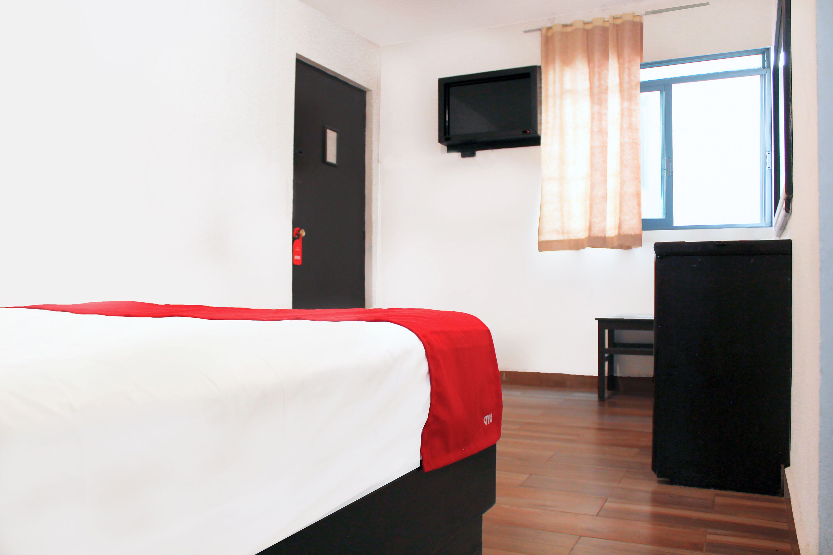 Quarto de hotel com cama, televisão na parede, escrivaninha e janela com cortinas.