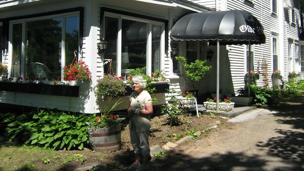 A house with a covered porch, large windows, and a garden with potted plants and flowers. - The Olde Village Inn (Sandy Cove)