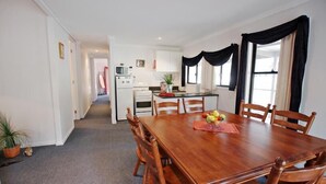 A dining area with a wooden table and chairs, a kitchen in the background, and a potted plant on the floor. - Serenity Grove (Fordwich)