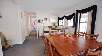 A dining area with a wooden table and chairs, a kitchen in the background, and a potted plant on the floor.