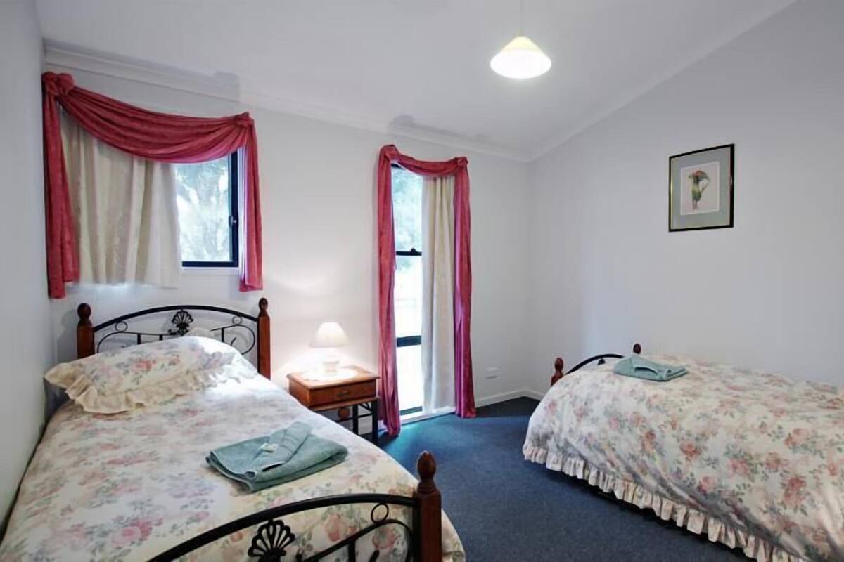 A bedroom with two single beds, a nightstand with a lamp, a window with curtains, and a framed picture on the wall.