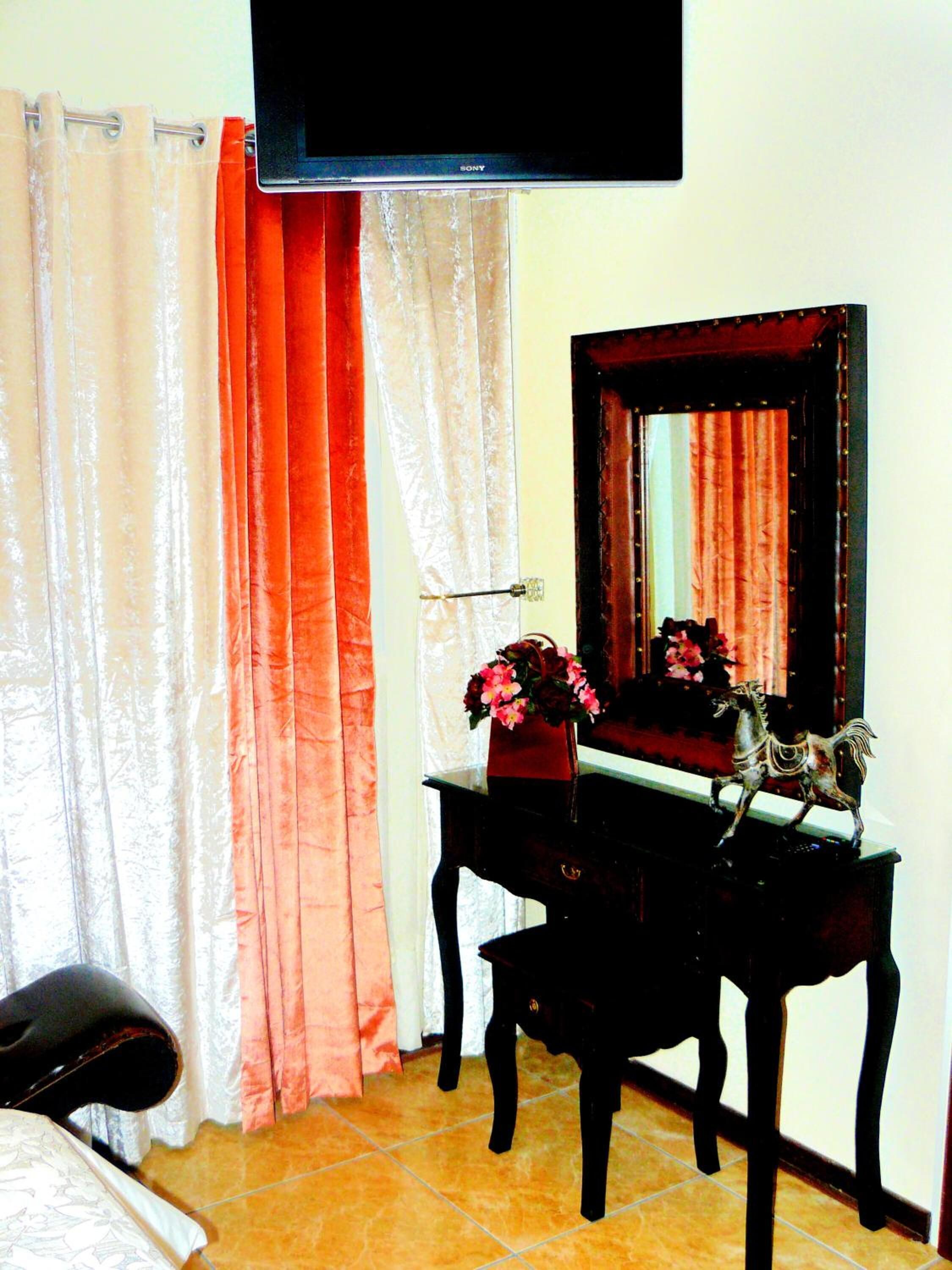A room with a TV mounted on the wall, a wooden dresser with a mirror, a vase with flowers, and a chair.