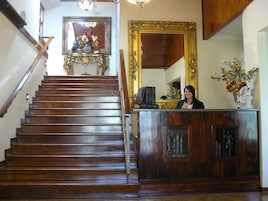 Uma escadaria de madeira com corrimão, uma mesa de recepção com um vaso de flores e um espelho grande com moldura ornamentada.