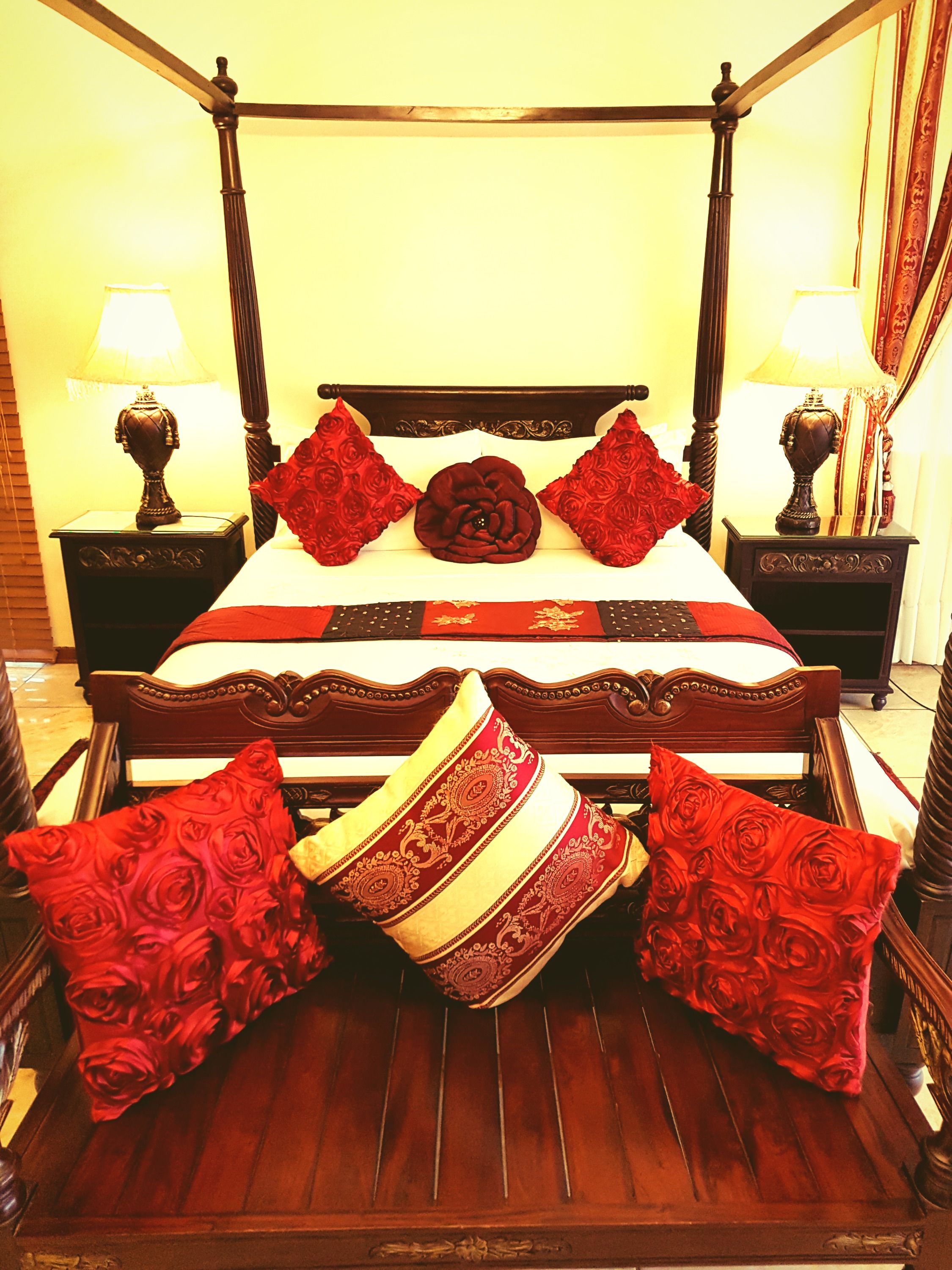 Una cama con dosel, con un cojín central con forma de rosa, flanqueado por dos cojines estampados con motivos de rosas, y una manta con estampado rojo y dorado.