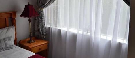 A bedroom with a bed, a nightstand with a lamp, a window with sheer curtains, and an air conditioning unit.