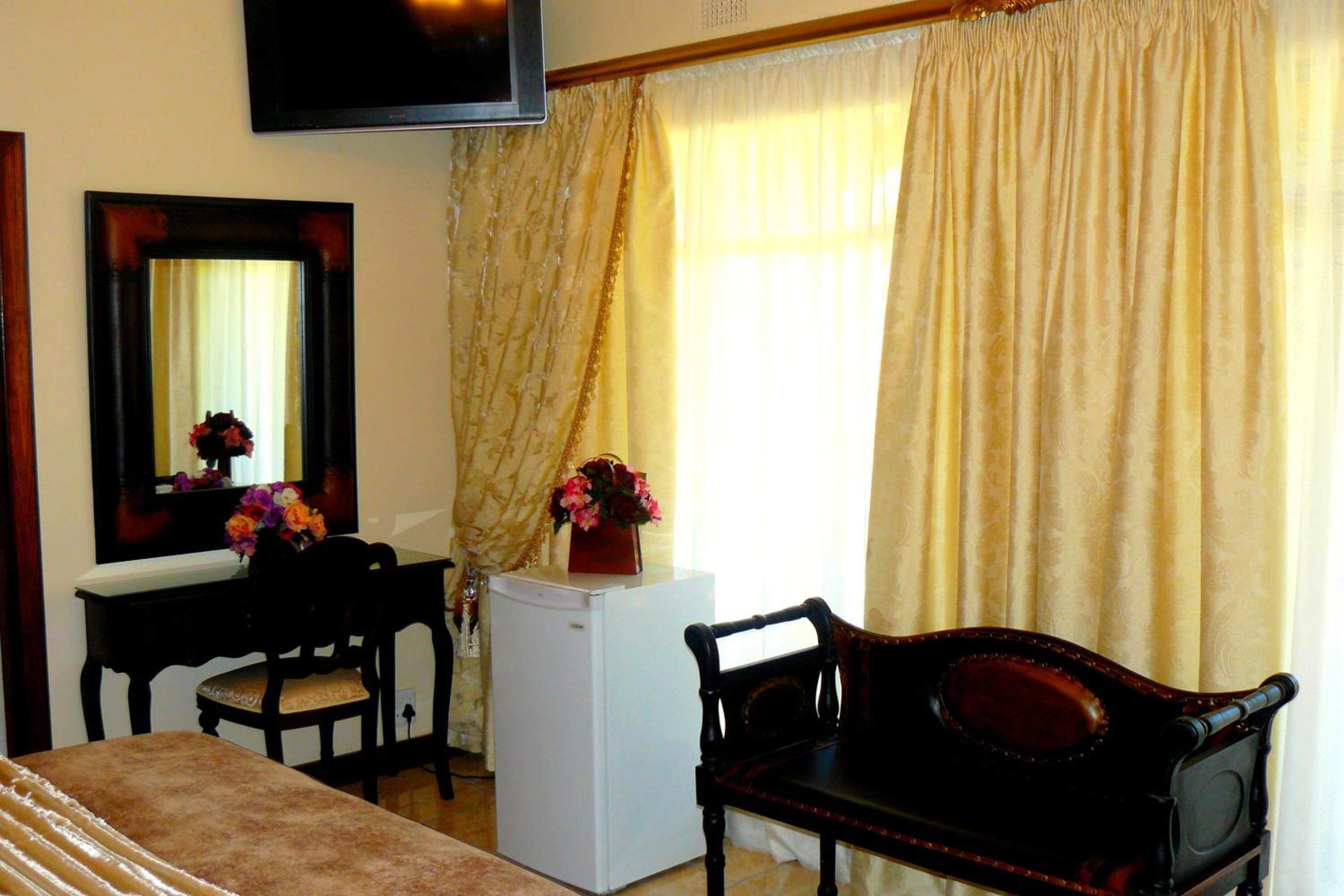 A room with a bed, a desk with a chair, a small white refrigerator, a mirror, and a television on the wall.