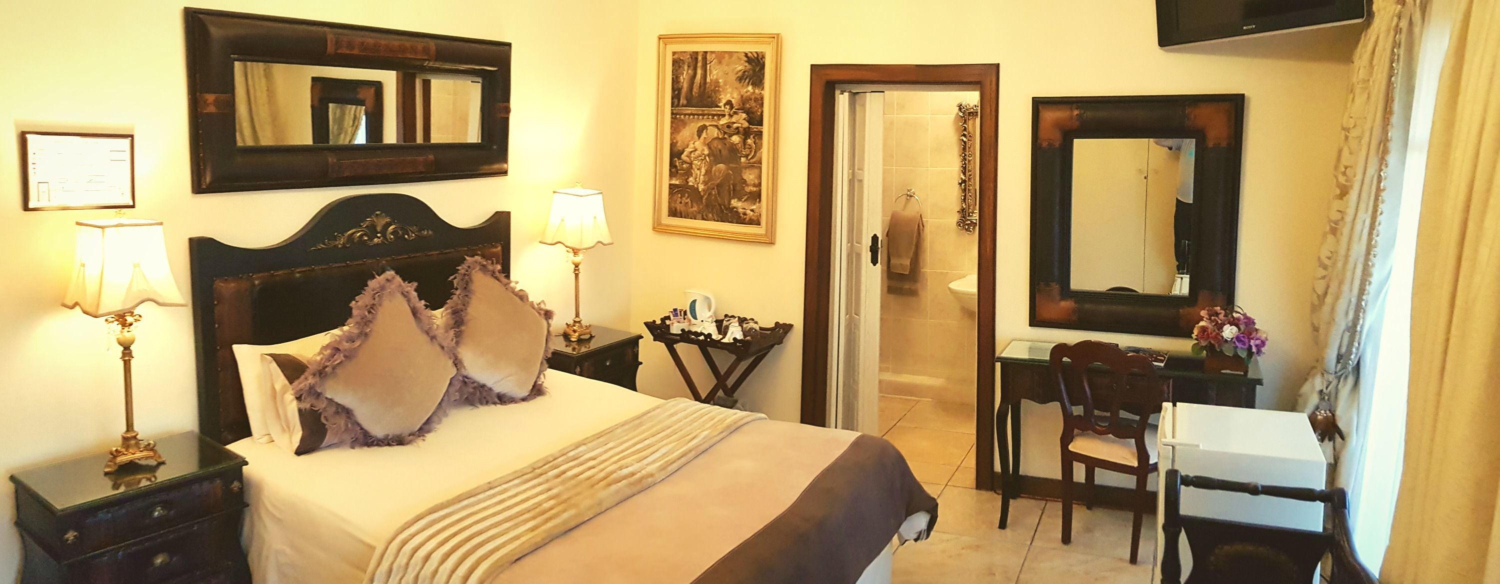 A hotel room with a bed, bedside tables, a desk, a chair, a mirror, and a door leading to a bathroom.