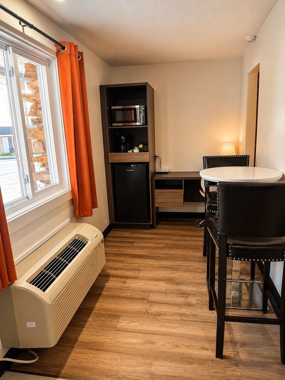 Une pièce compacte dotée d’une fenêtre, d’un climatiseur intégré, d’un four à micro-ondes, d’un réfrigérateur, d’une table à manger avec des chaises et d’une lampe.