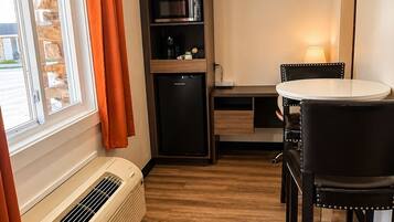 Une pièce compacte dotée d’une fenêtre, d’un climatiseur intégré, d’un four à micro-ondes, d’un réfrigérateur, d’une table à manger avec des chaises et d’une lampe.