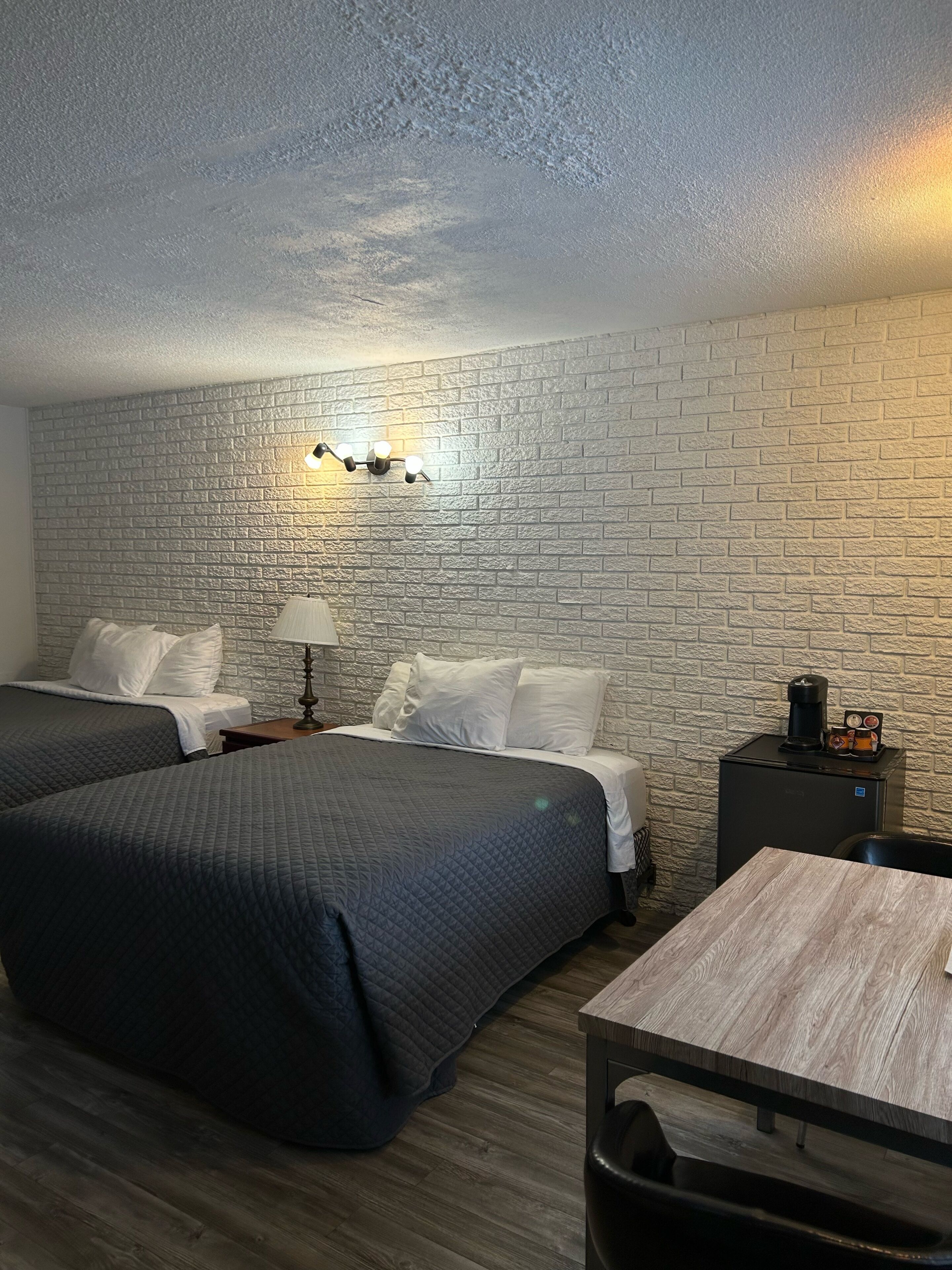 A hotel room with two beds, a small table, a lamp, a coffee maker, and a trash bin.