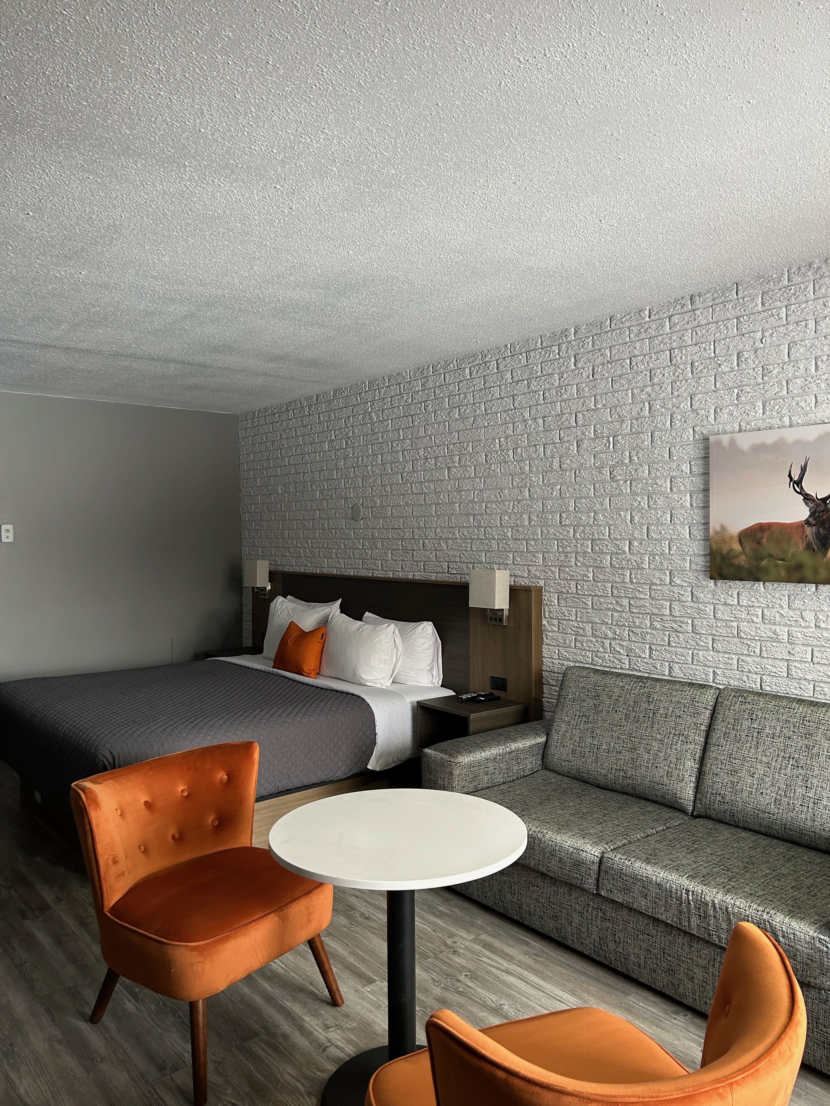 A modern hotel room with a brick-patterned wall, a large bed with white and orange pillows, a grey sofa, and two orange chairs around a small white table.