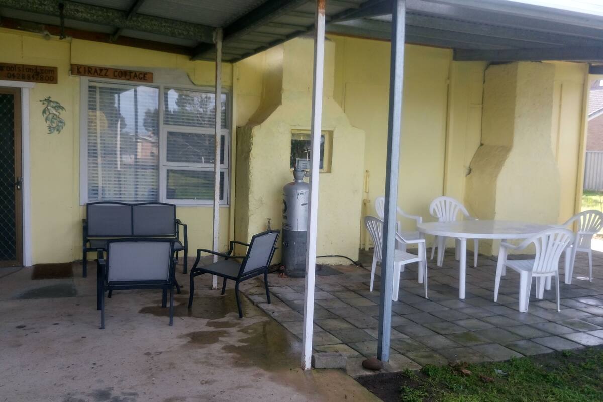 Un área de estar al aire libre cubierta, con sillas y una mesa, un cilindro de gas y una puerta con un letrero encima.