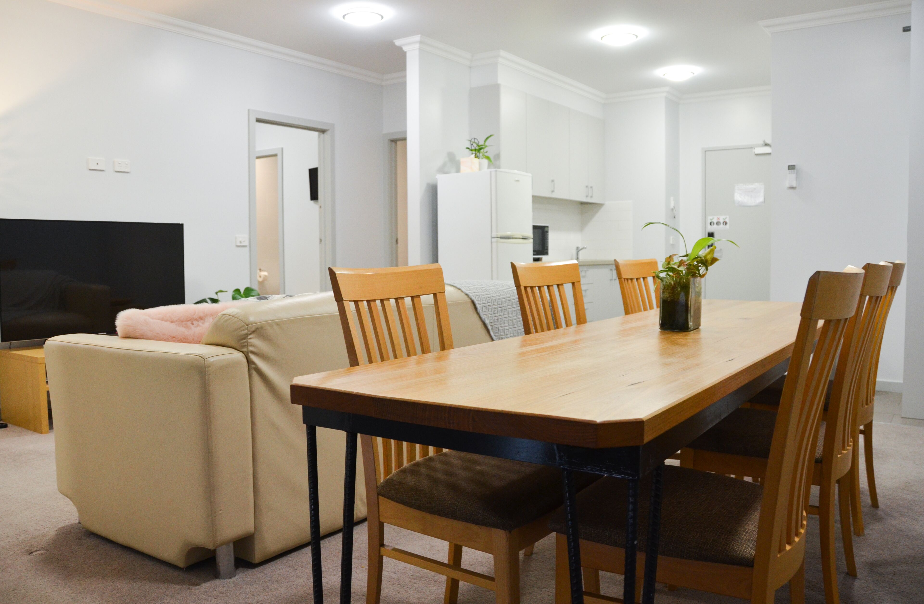 A modern living room with a beige sofa, a wooden dining table with six chairs, a flat-screen TV, and a kitchen area in the background.
