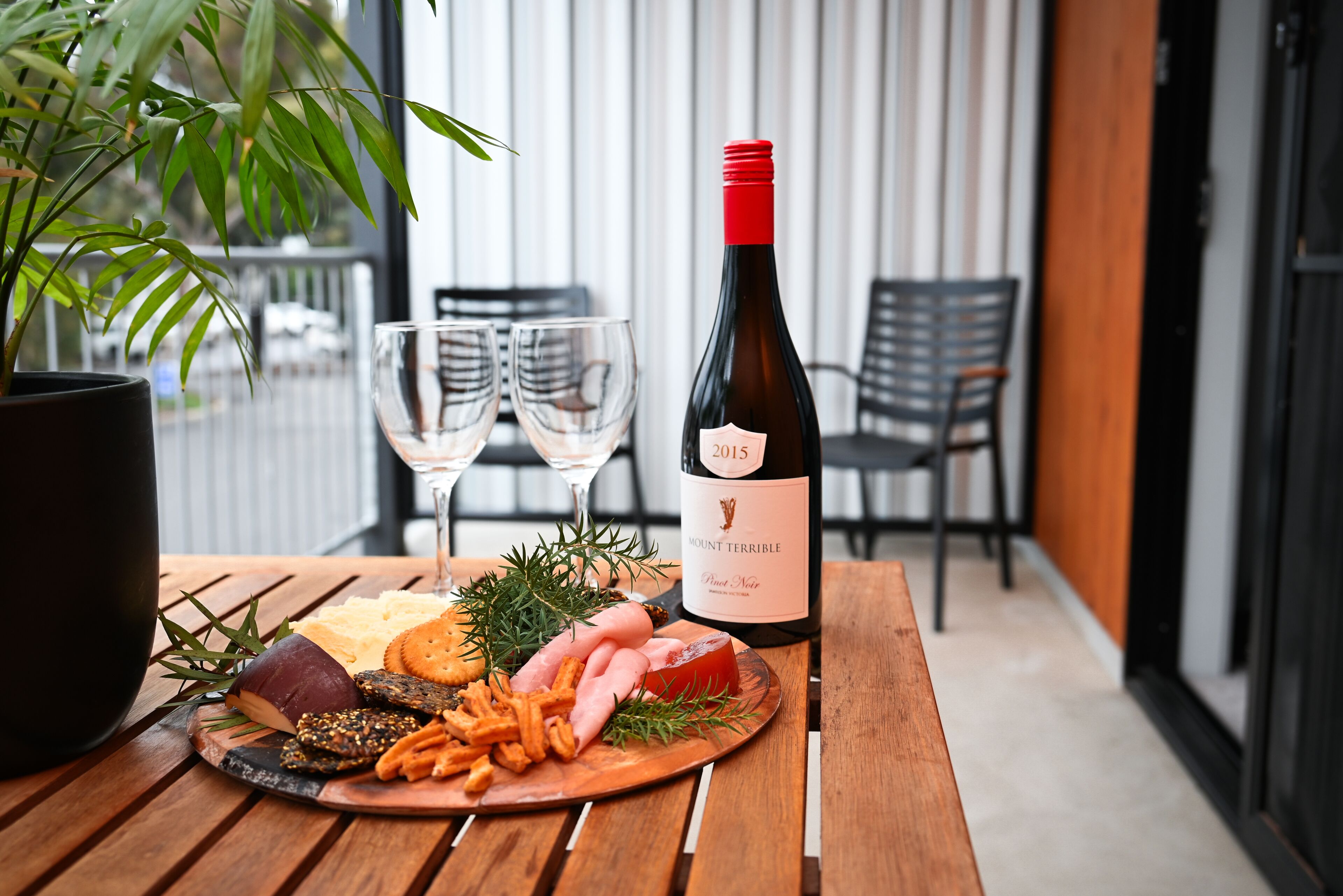 A wooden table with a charcuterie board, two wine glasses, and a bottle of wine on a balcony with chairs and a potted plant.