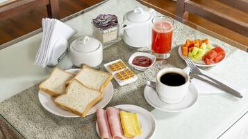 Ein Frühstückstisch mit Brot, Schinken, Käse, einem Teekessel, einer Tasse Kaffee, einem Glas Saft und einer Schale Obst.