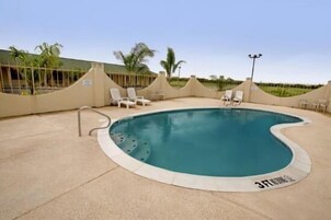 Piscina com borda ondulada, rodeada por um deck bege e cadeiras de descanso brancas.
