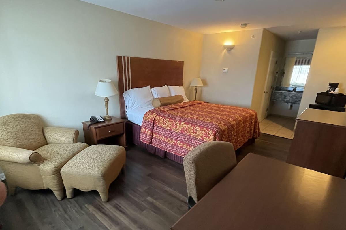 A hotel room with a bed, a chair, a table, a nightstand, and a lamp.
