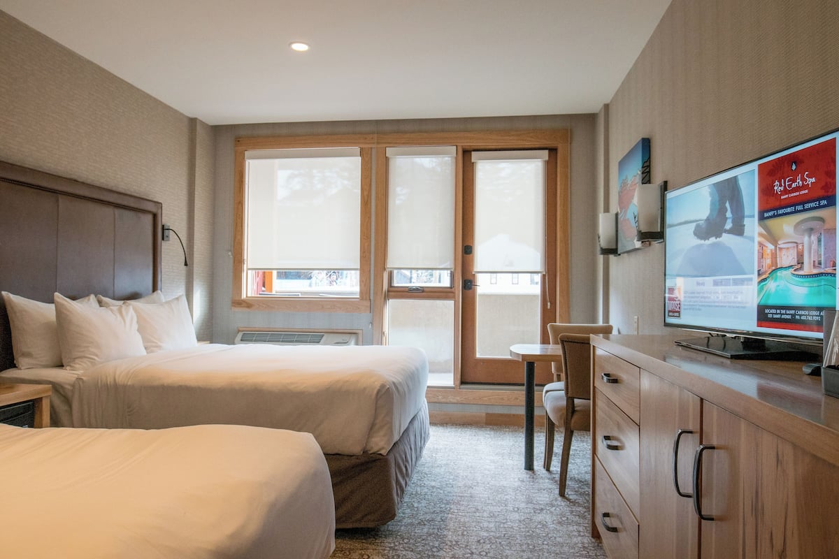 Habitación de hotel con dos camas, un escritorio, televisión y vista al exterior mediante una puerta corrediza.