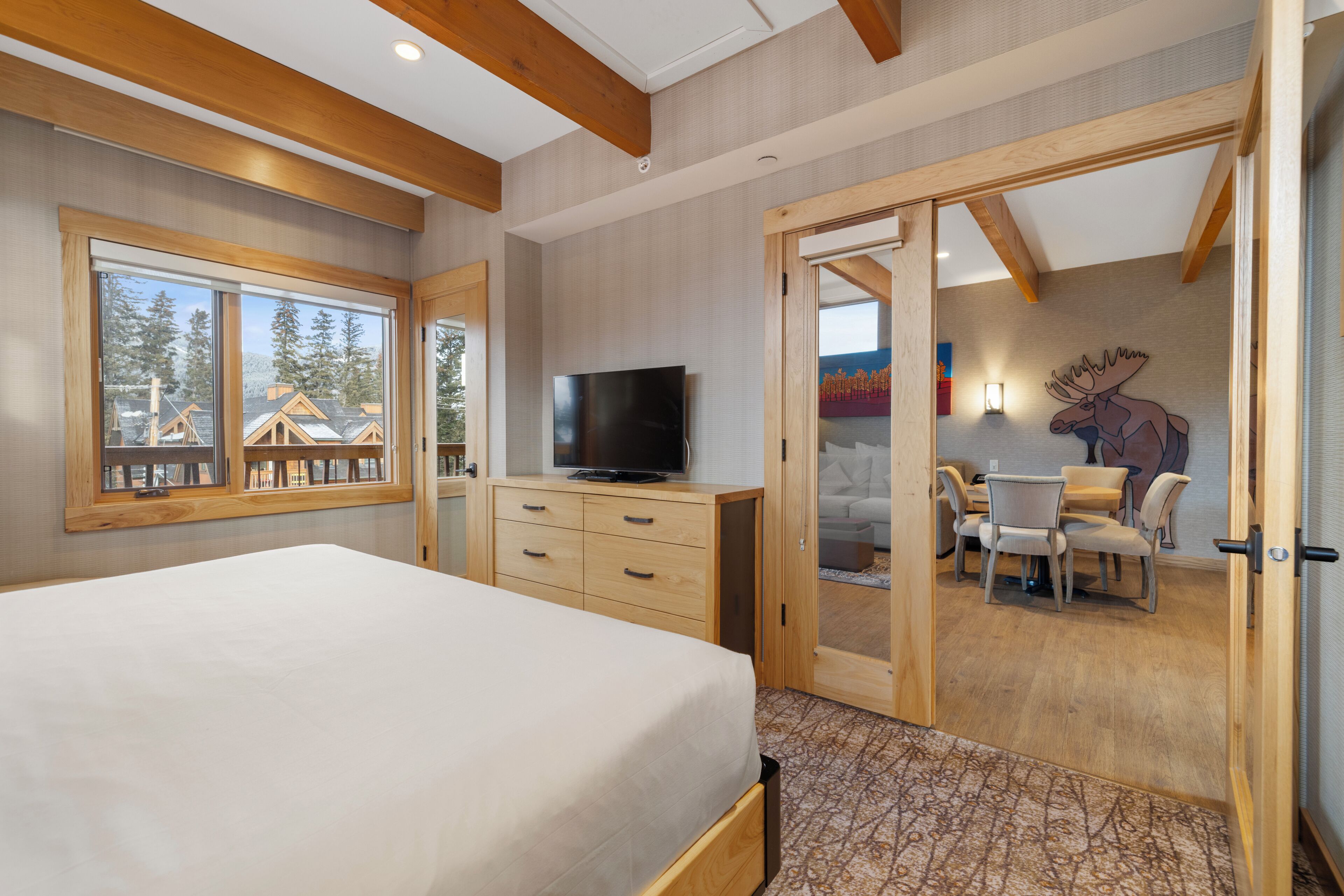A bedroom with a bed, a TV, a wooden dresser, and a sliding door leading to a dining area with a table and chairs.