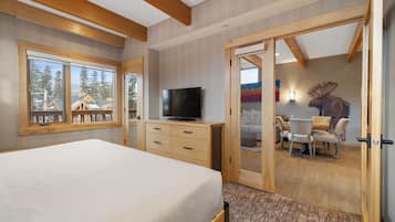 A bedroom with a bed, a TV, a wooden dresser, and a sliding door leading to a dining area with a table and chairs.