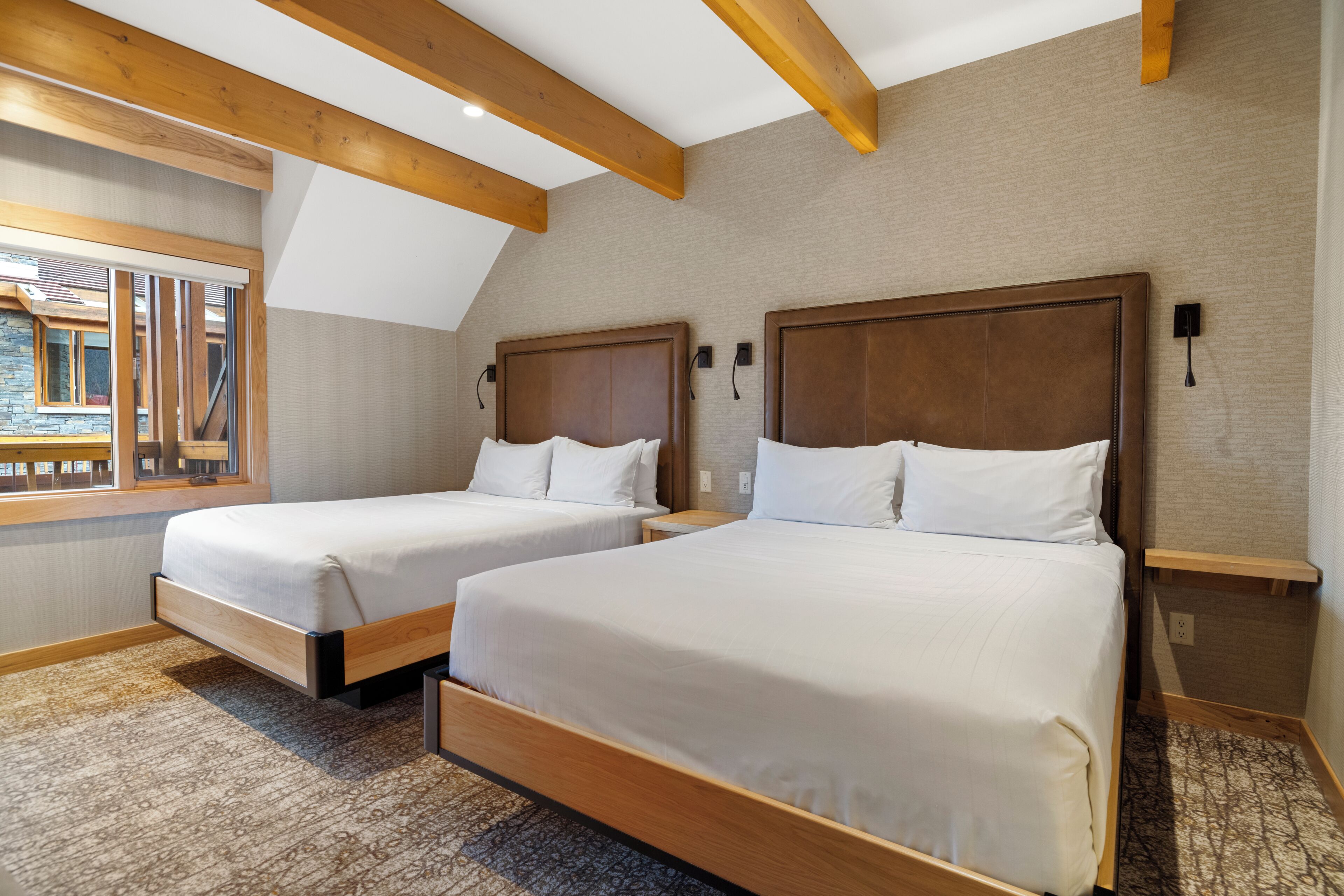A hotel room with two beds, wooden beams on the ceiling, a window with a view, and a carpeted floor.