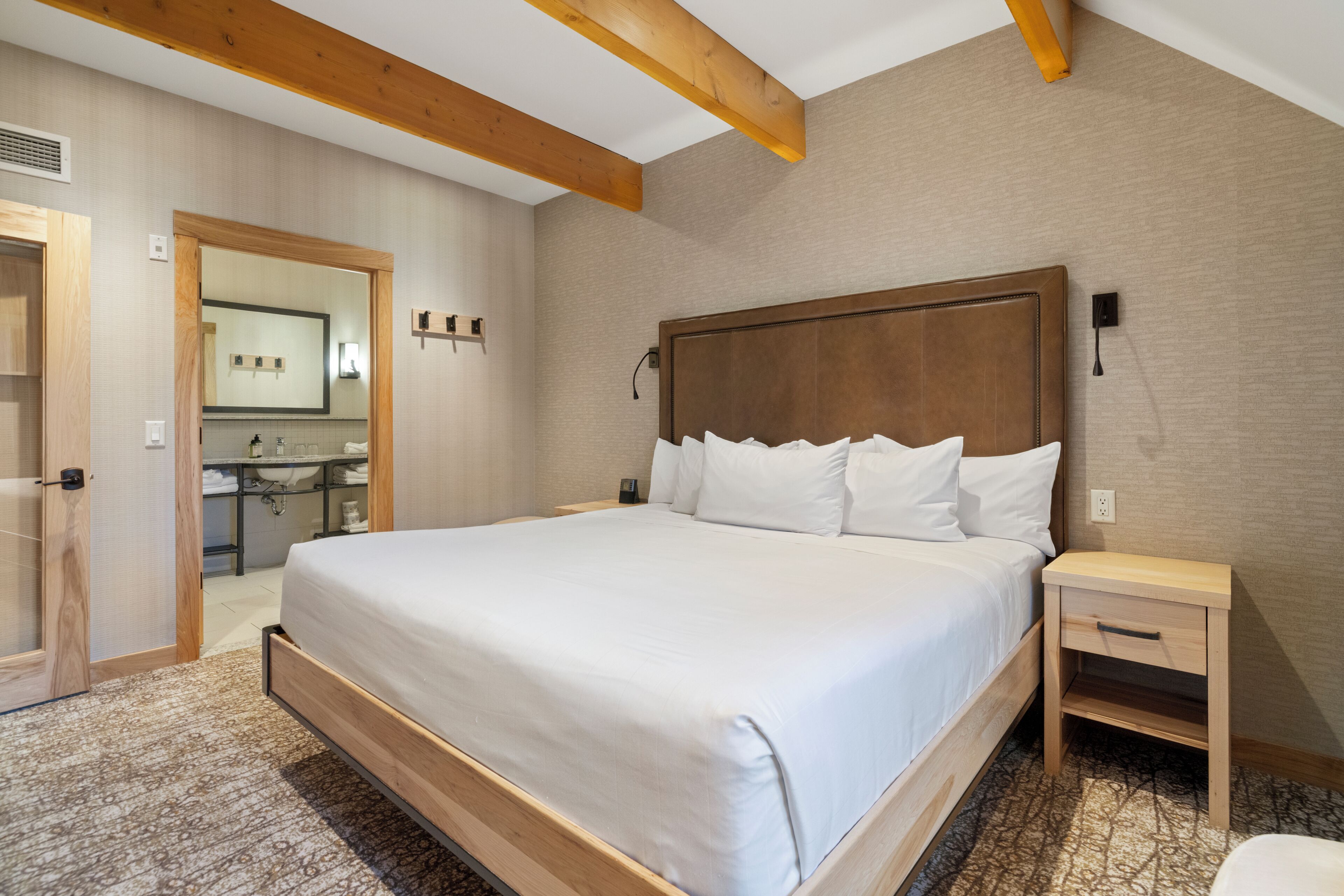 A hotel room with a large bed, wooden headboard, a bedside table with a drawer, and a bathroom visible through an open door.