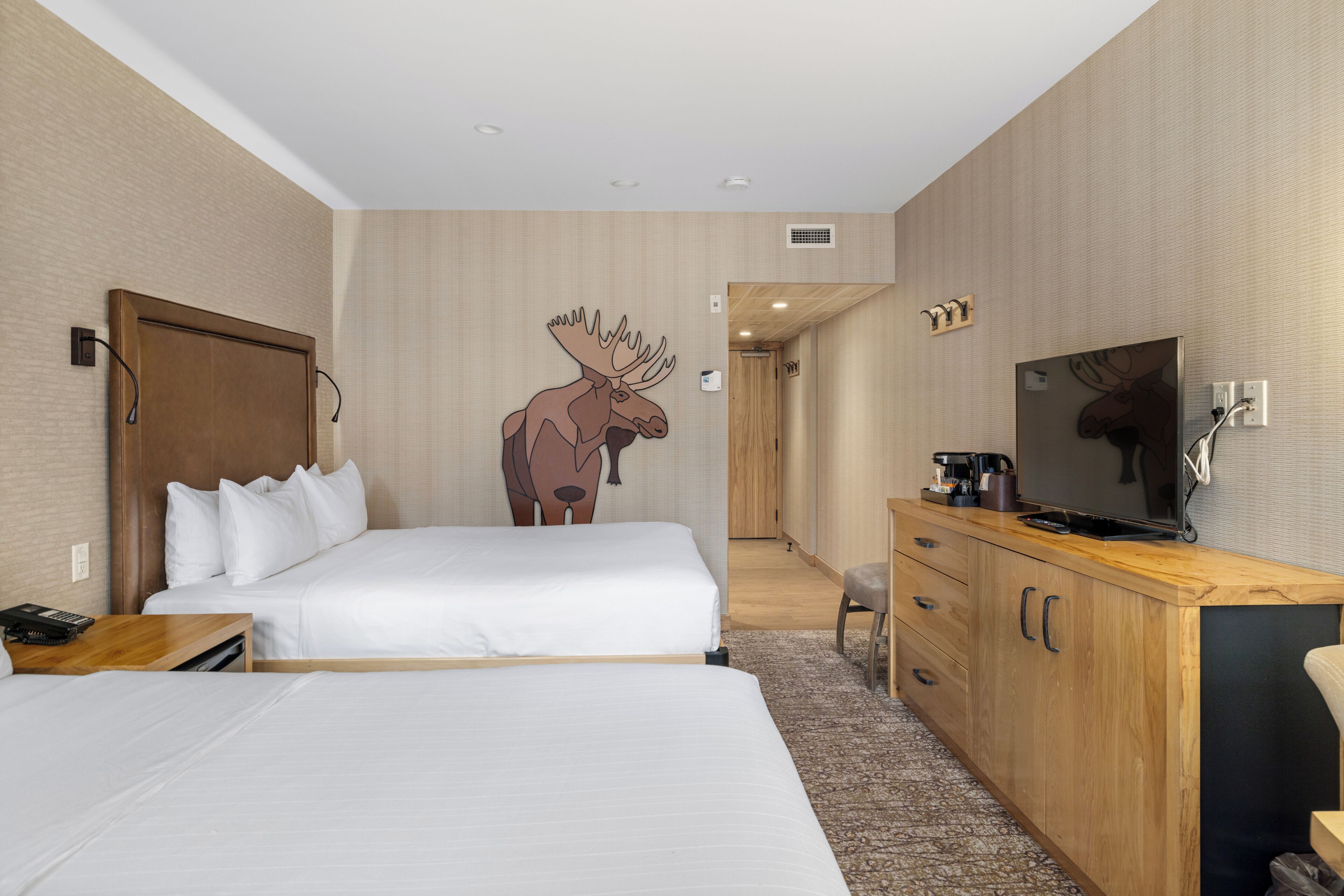 A hotel room with two beds, a wooden dresser, a television, and a moose wall decal.