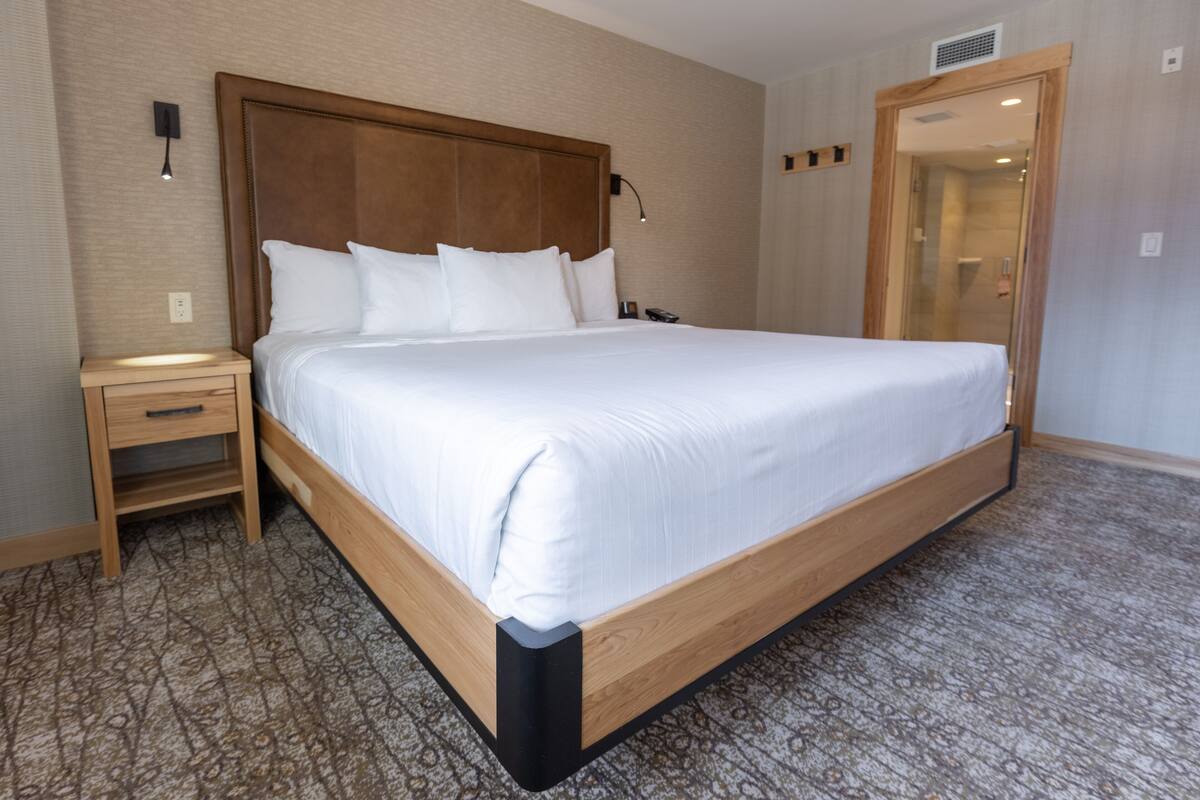 Un lit bien fait, avec du linge de lit blanc, dans une chambre d’hôtel dotée d’une tête de lit en bois, d’une table de chevet et d’un tapis à motifs.