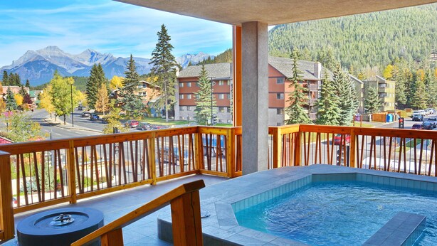 Een balkon met een jacuzzi, houten reling en uitzicht op een berglandschap en gebouwen.