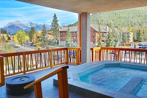 Een balkon met een jacuzzi, houten reling en uitzicht op een berglandschap en gebouwen.