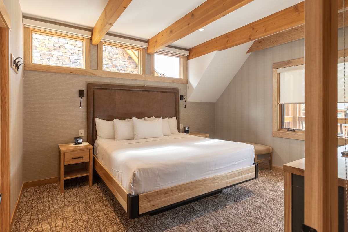 Une chambre à coucher avec un grand lit, une tête de lit en bois, deux tables de chevet, un banc et une fenêtre donnant sur un mur en pierre.