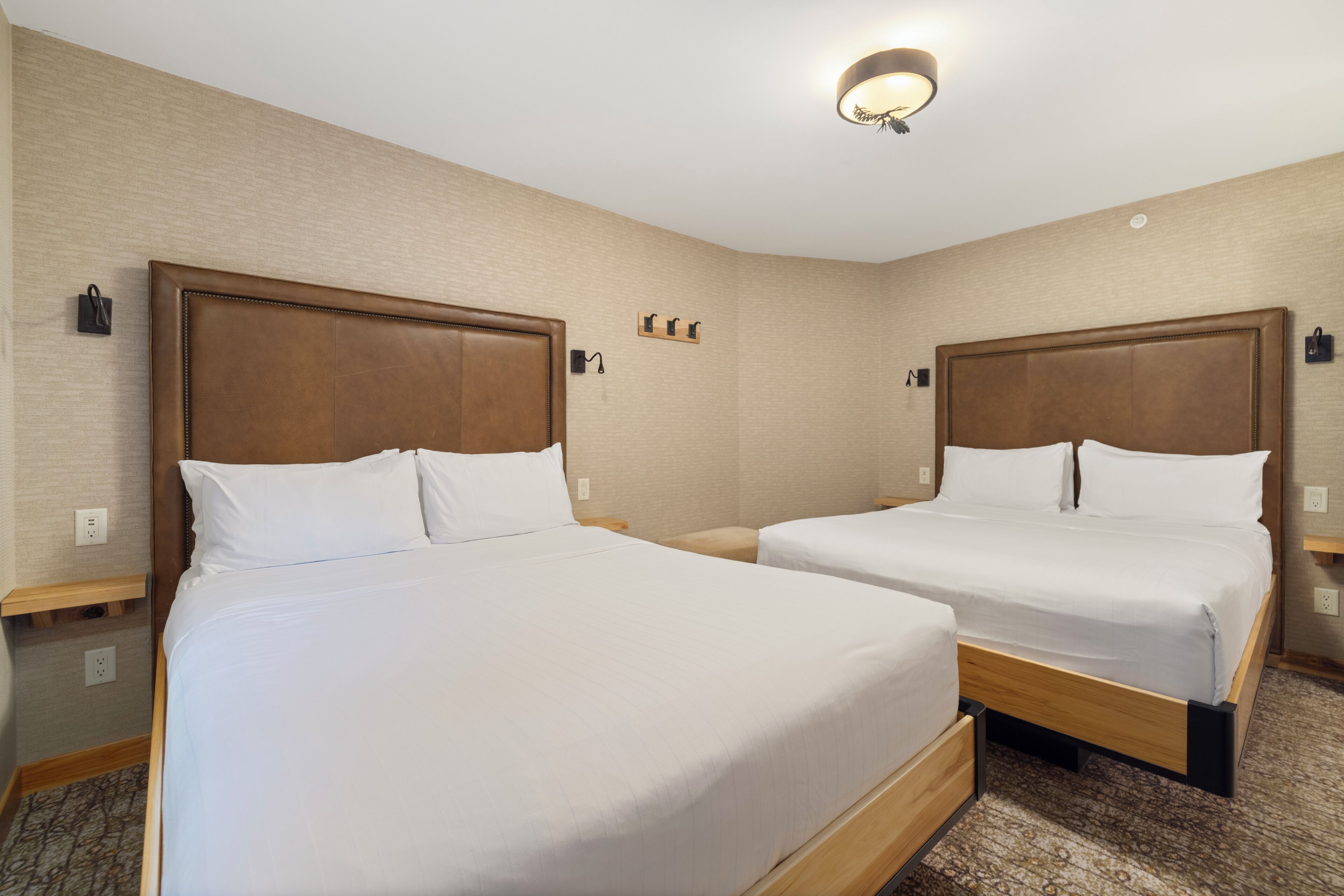 A hotel room with two beds, a wooden headboard, a nightstand, a wall-mounted lamp, and a carpeted floor.