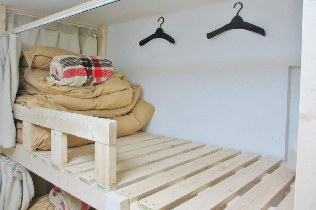 Quarto pequeno com uma cama de solteiro, estrutura de madeira, janela com cortinas e dois cabides pretos na parede.