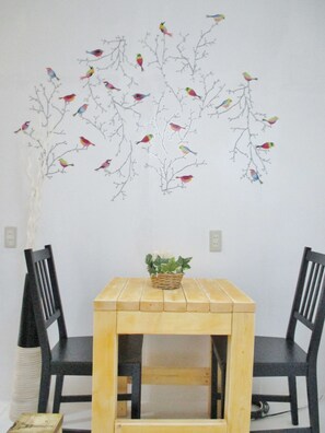 A dining area with a wooden table and two black chairs. There is a wall decoration of colorful birds on branches. - Hostel AI (Osaka)