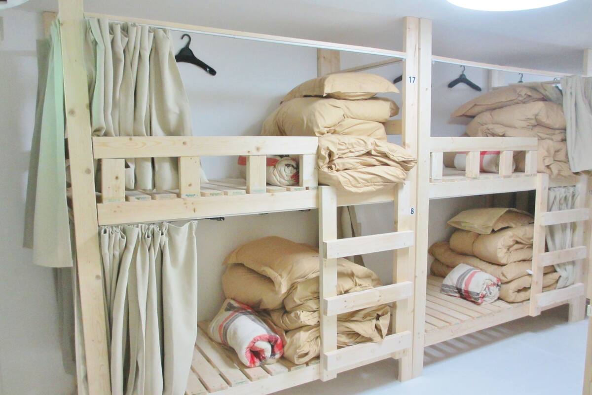 Quarto com beliches de estrutura de madeira, roupa de cama bege e cortina clara.