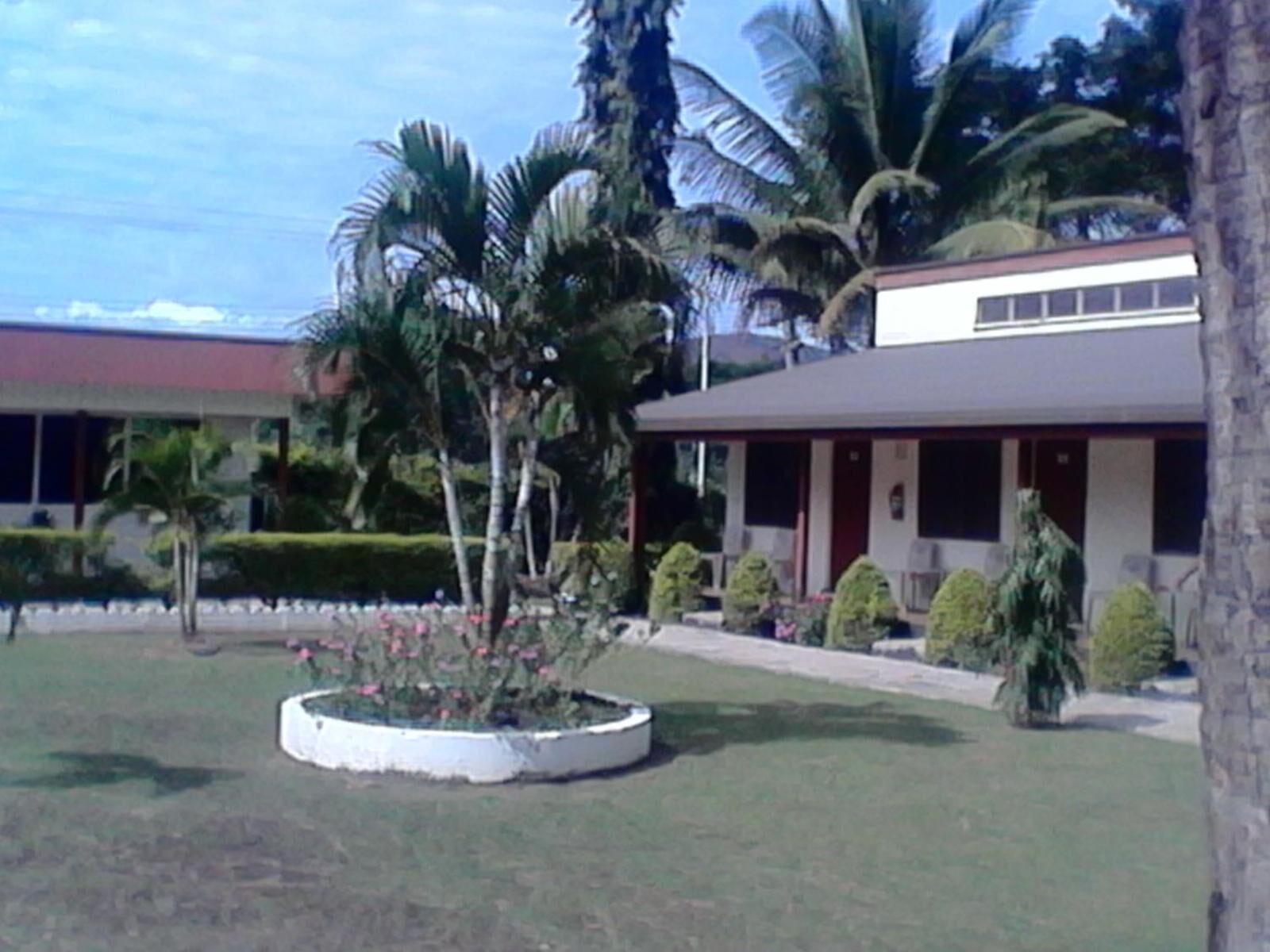 A building with a covered porch, palm trees, and a landscaped garden with a circular flower bed.