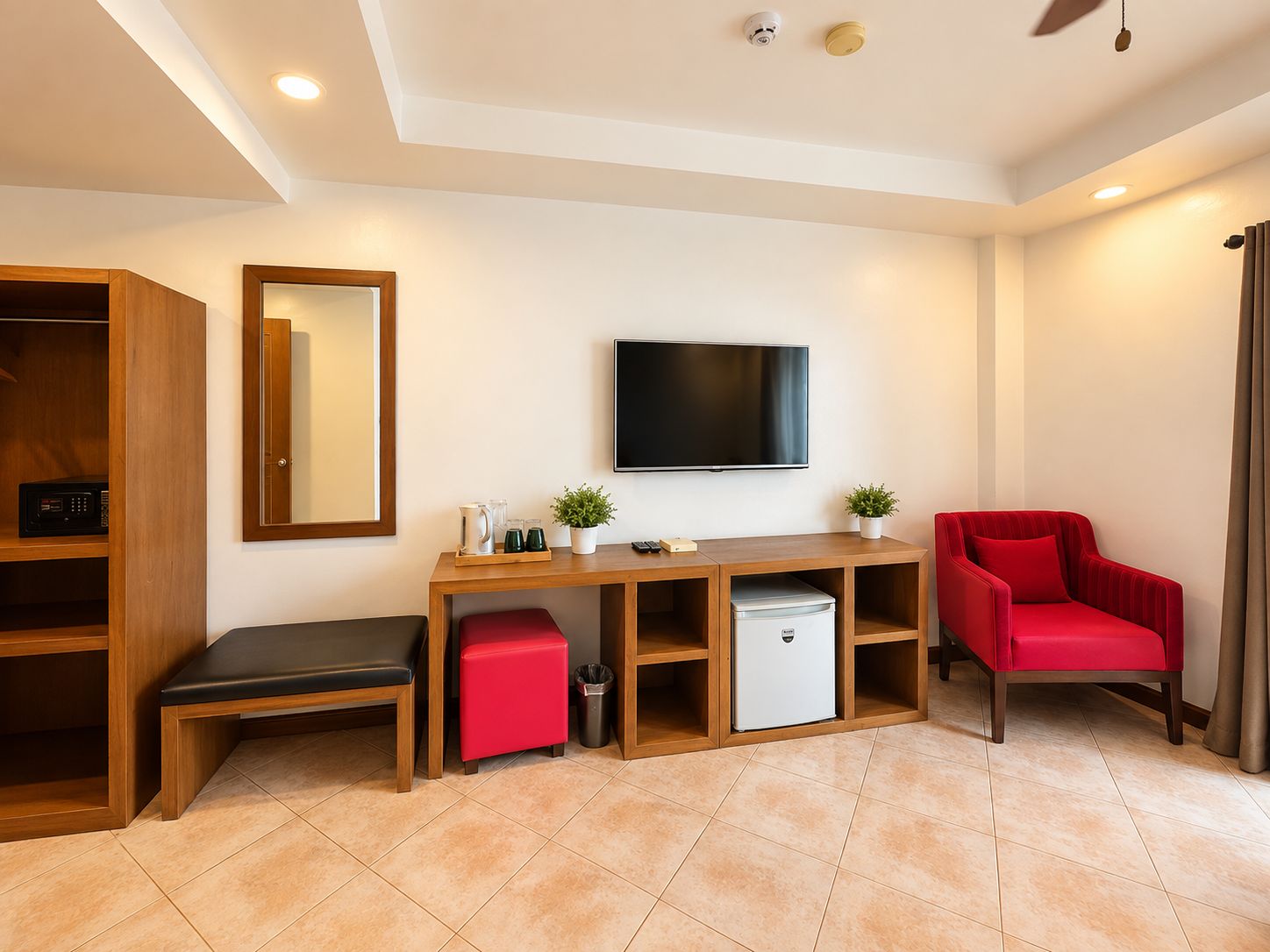 A hotel room with a wooden desk, a television, a red chair, a small refrigerator, and a trash bin.