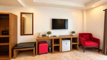 A hotel room with a wooden desk, a television, a red chair, a small refrigerator, and a trash bin.